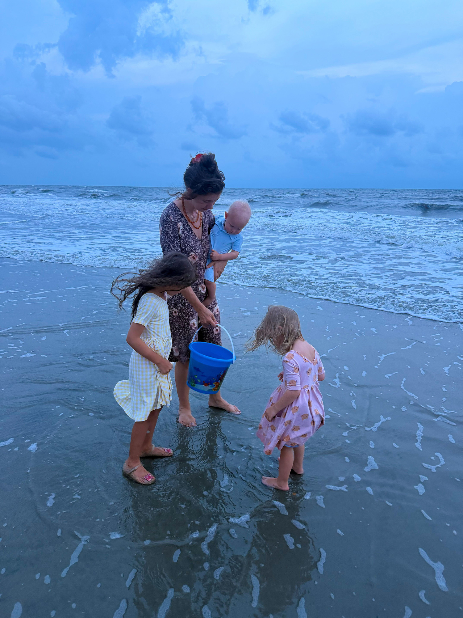 A woman in a patterned dress stands in shallow ocean water holding a baby while two young girls in light dresses play with a blue bucket at the cloudy beach.