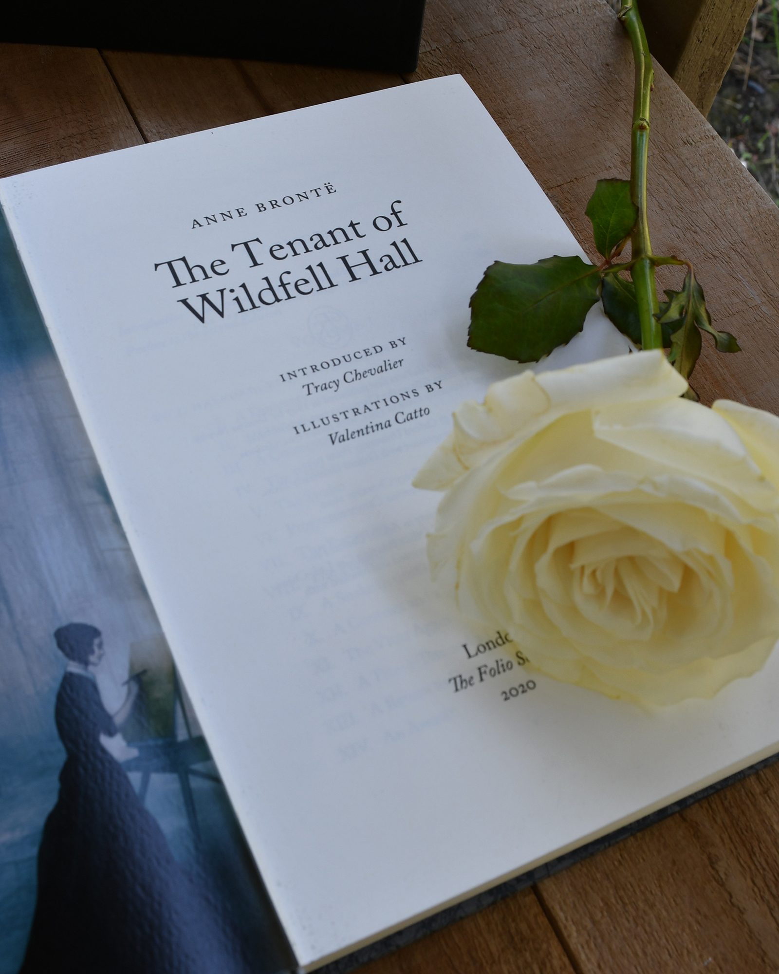 Open book page showing The Tenant of Wildfell Hall title with a white rose laid across the page on a wooden surface.